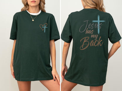 May include: Dark green t-shirt with a white collar and a gold necklace. The front of the shirt features a heart and cross design. The back of the shirt has the text "Jesus has my Back" with a cross graphic.