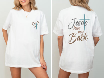 May include: White t-shirt with a heart and cross design on the front and the text "Jesus has my Back" on the back. The design features a teal cross and brown heart and text. The shirt is a short-sleeved, casual style.