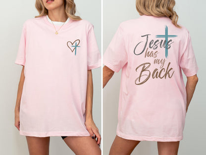 May include: A light pink t-shirt with a heart and cross design on the front. The back of the shirt features the text "Jesus has my Back" with a cross graphic. The shirt is a comfortable, casual style.