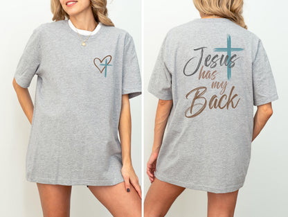 May include: A light gray t-shirt with short sleeves. The front features a heart and cross design. The back of the shirt displays the text "Jesus has my Back" with a cross graphic. The shirt is made of a soft material.
