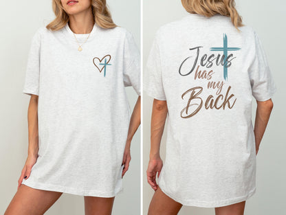 May include: Light gray t-shirt with a heart and cross design on the front and the text "Jesus has my Back" on the back. The design features a teal cross and brown text. The shirt is a casual, oversized fit.