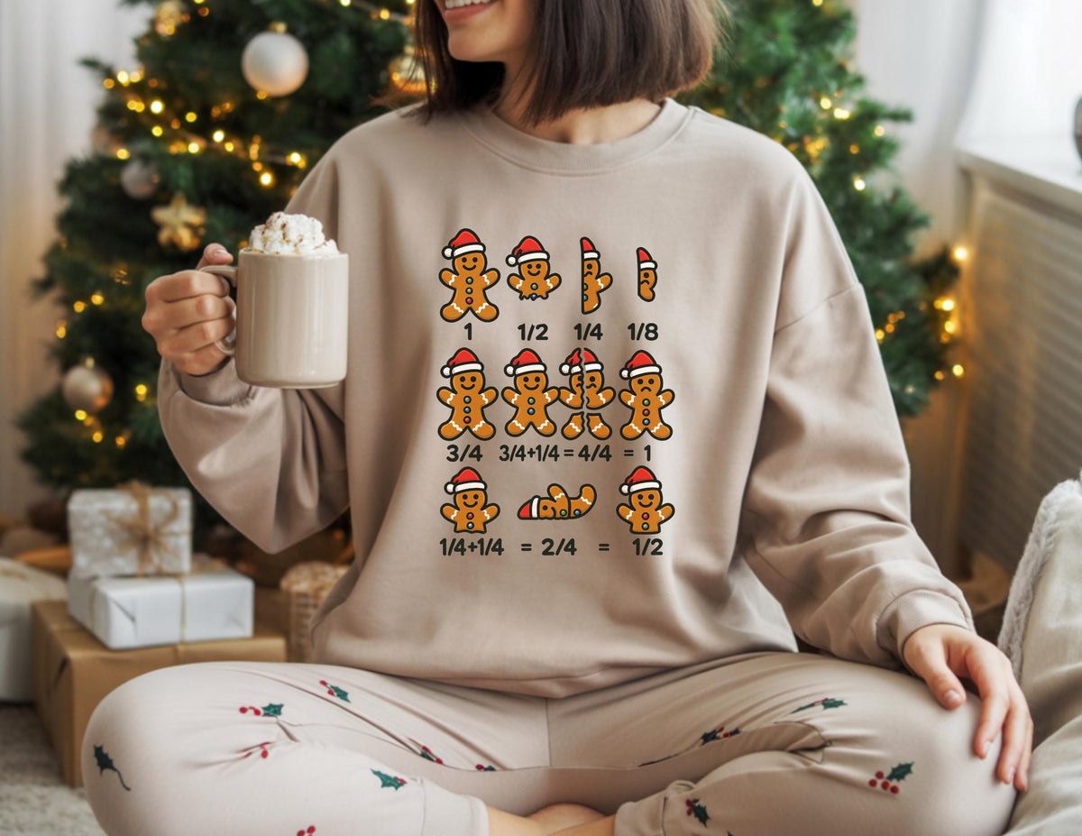May include: Beige sweatshirt with a gingerbread man math graphic. The graphic shows gingerbread men in Santa hats with fractions. The person is holding a beige mug with whipped cream. The person is wearing matching pajama pants with Christmas holly.
