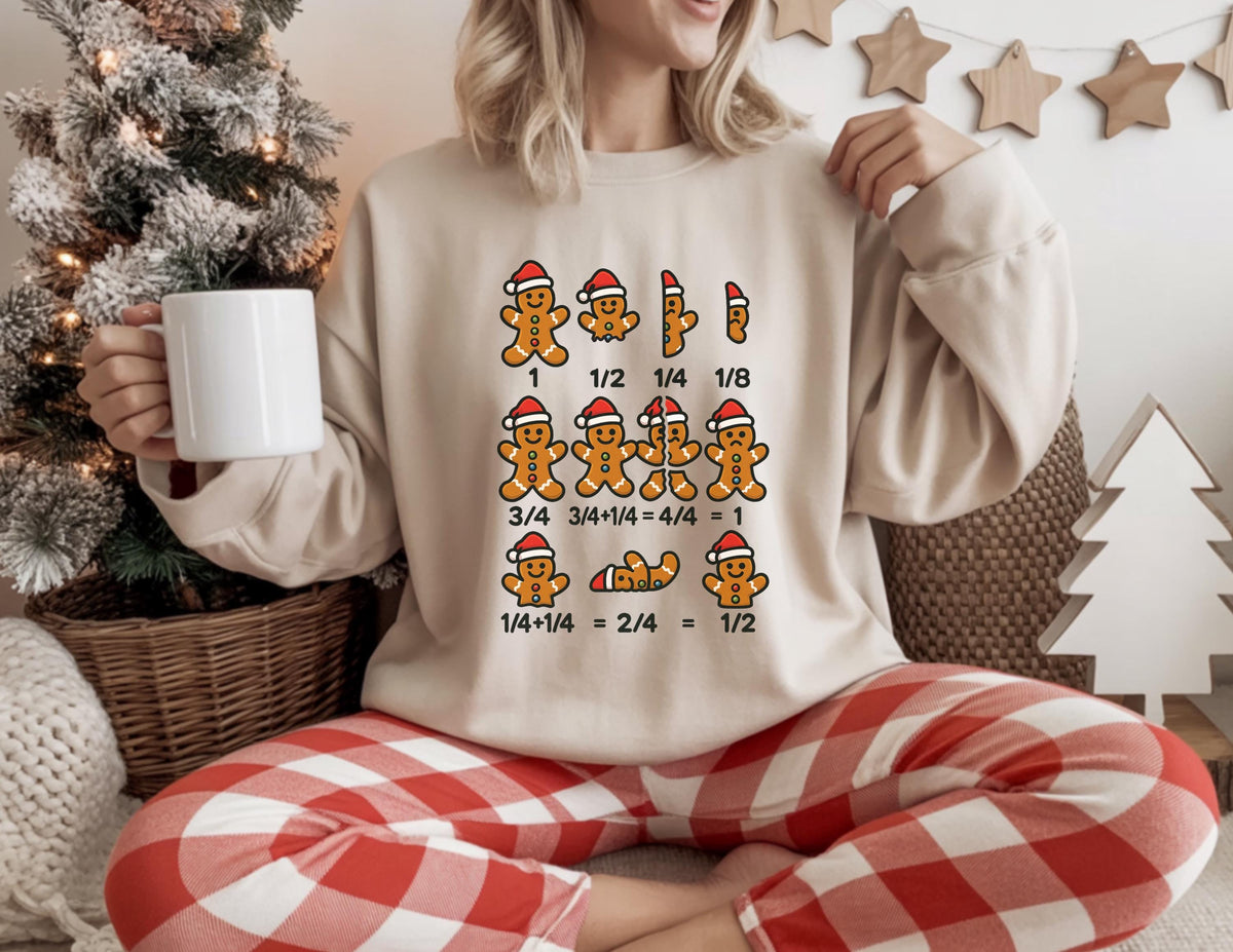 May include: Beige sweatshirt with a gingerbread man graphic wearing a Santa hat. The graphic shows fractions with gingerbread men. The person is wearing red and white plaid pants and holding a white mug. A Christmas tree and decorations are in the background.