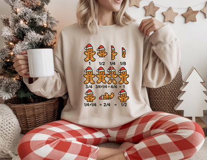 May include: Beige sweatshirt with a gingerbread man graphic wearing a Santa hat. The graphic shows fractions with gingerbread men. The person is wearing red and white plaid pants and holding a white mug. A Christmas tree and decorations are in the background.