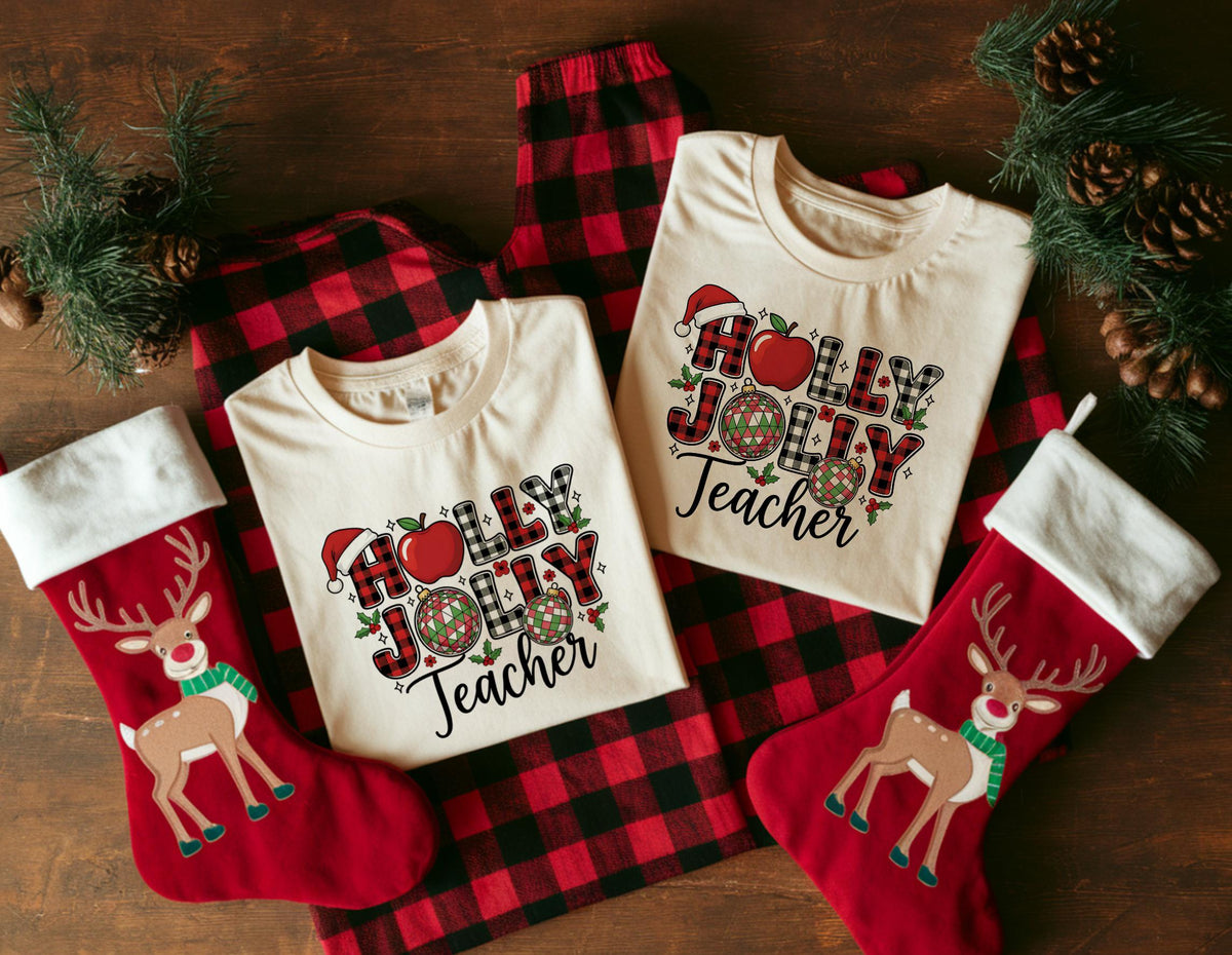 May include: Two cream-colored t-shirts with the text "HOLLY JOLLY Teacher" and Christmas graphics, including a Santa hat and apple. The shirts are on a red and black plaid pajama set, with red Christmas stockings and pine branches.