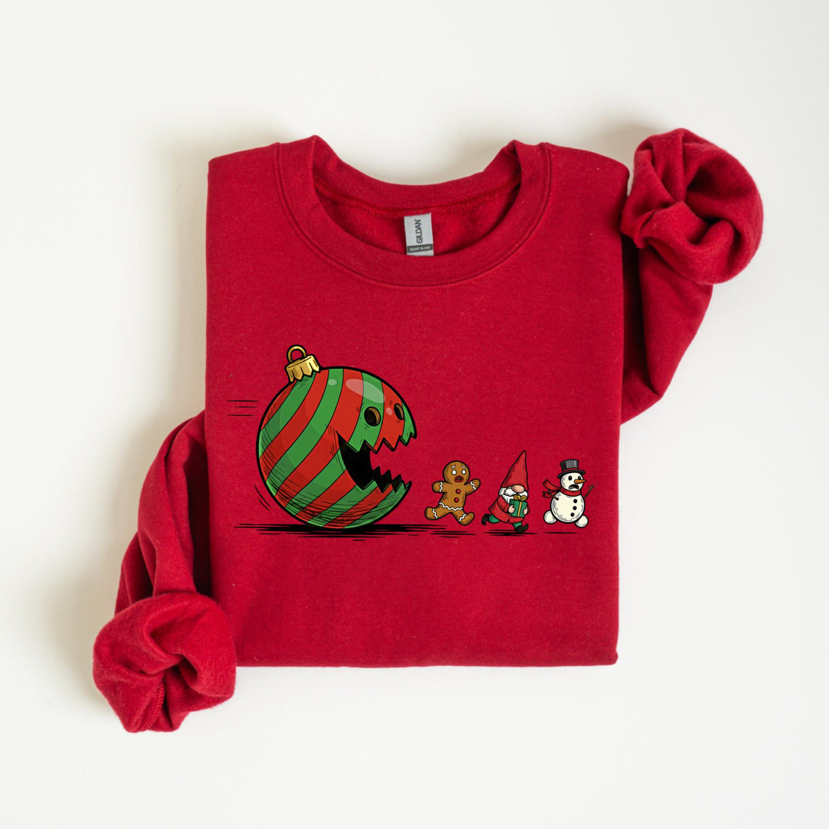 May include: A red sweatshirt featuring a Christmas ornament with a bite taken out of it, with green and red stripes. A gingerbread man, gnome, and snowman are walking to the right. The sweatshirt is a solid red color.