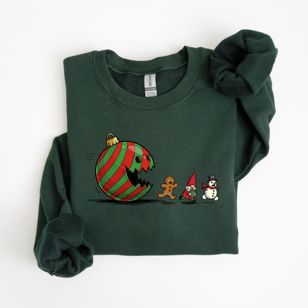 May include: Dark green sweatshirt featuring a whimsical Christmas ornament design. The ornament, with red and green stripes, appears to be eating a gingerbread man, a gnome, and a snowman. The sweatshirt is a crew neck style.