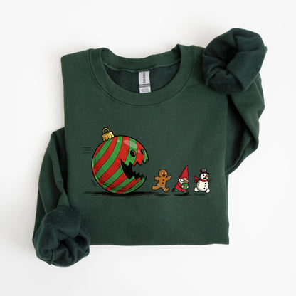 May include: Dark green sweatshirt featuring a whimsical Christmas ornament design. The ornament, with red and green stripes, appears to be eating a gingerbread man, a gnome, and a snowman. The sweatshirt is a crew neck style.