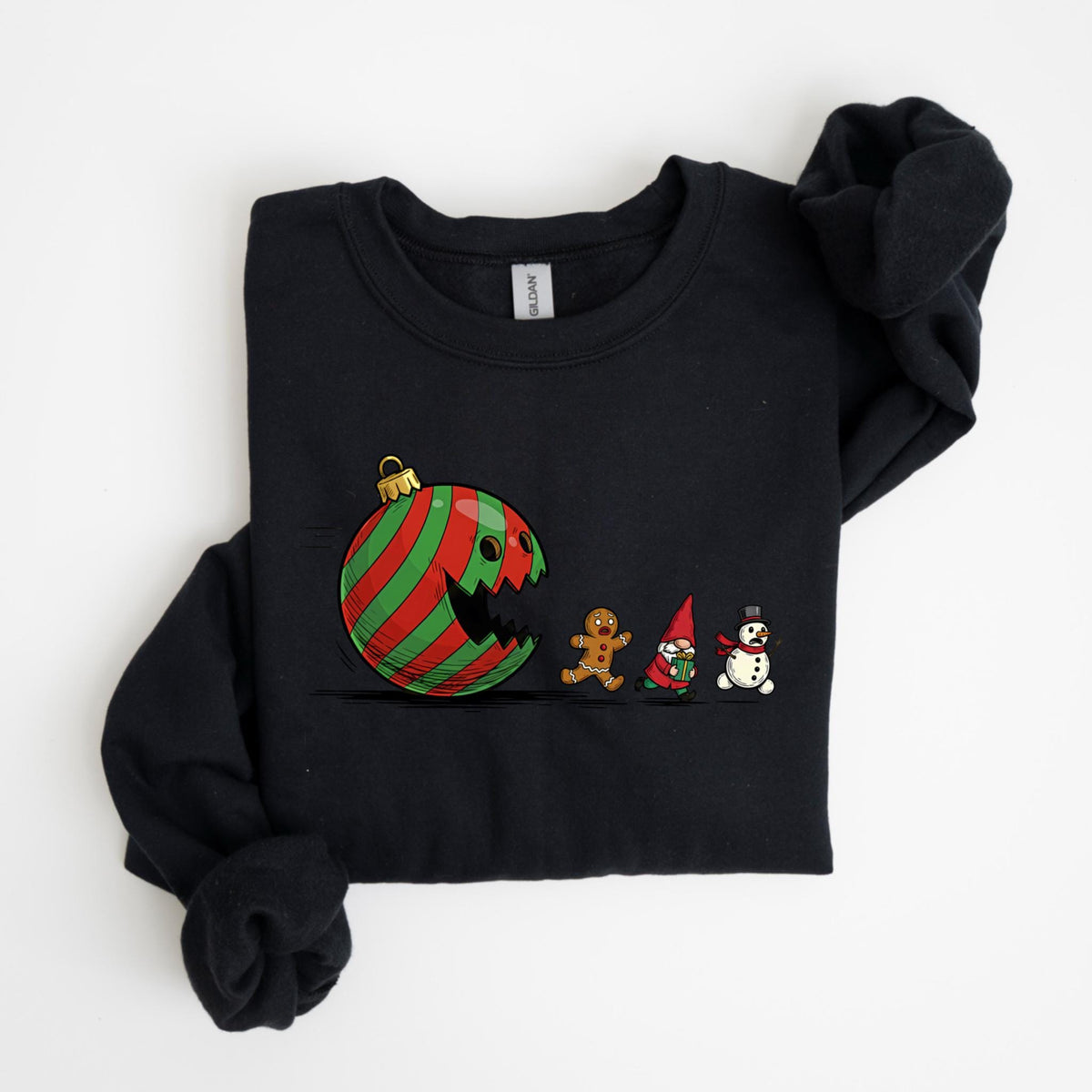 May include: Black sweatshirt featuring a Christmas ornament with a face, red and green stripes, and a bite taken out of it. A gingerbread man, gnome, and snowman are walking in a line. The sweatshirt is folded.