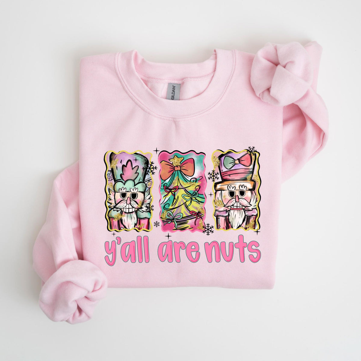 May include: A light pink sweatshirt featuring a colorful graphic of three nutcrackers and the text "y'all are nuts" in pink. The nutcrackers are in a watercolor style with pastel colors and festive details, perfect for the holiday season.