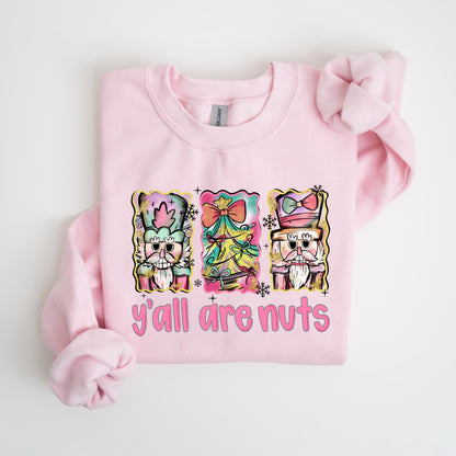 May include: A light pink sweatshirt featuring a colorful graphic of three nutcrackers and the text "y'all are nuts" in pink. The nutcrackers are in a watercolor style with pastel colors and festive details, perfect for the holiday season.