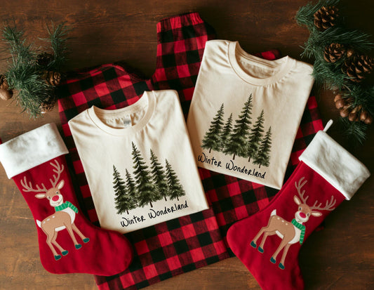 May include: A festive holiday scene featuring two cream-colored t-shirts with a forest graphic and the words "Winter Wonderland." The shirts are laid on red and black plaid pajama pants. Two red Christmas stockings with reindeer designs complete the cozy arrangement.
