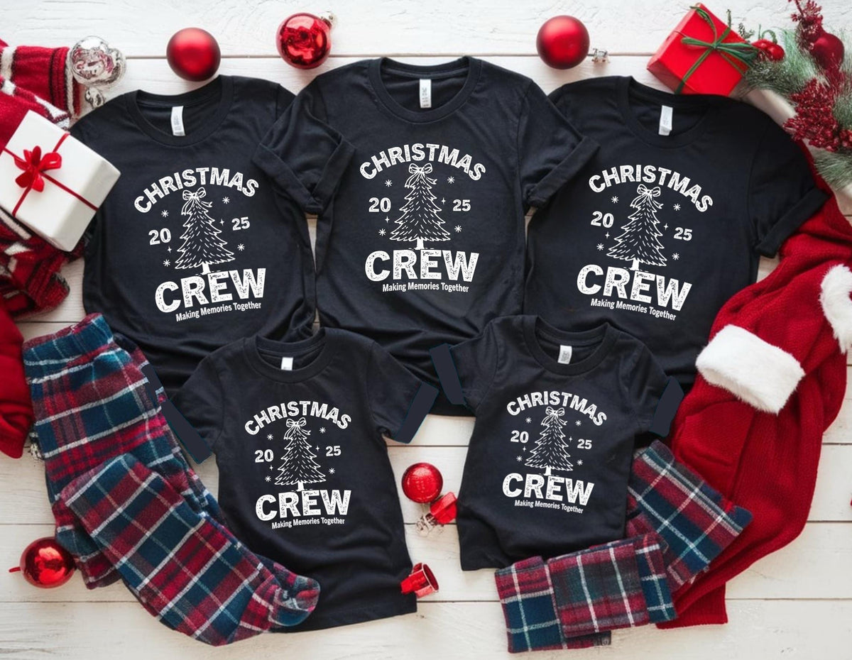 May include: Black t-shirts with a Christmas tree graphic and the text "CHRISTMAS CREW 2025 Making Memories Together." Red and blue plaid pajama pants and Christmas decorations surround the shirts. A white gift box and red Santa hat are also present.