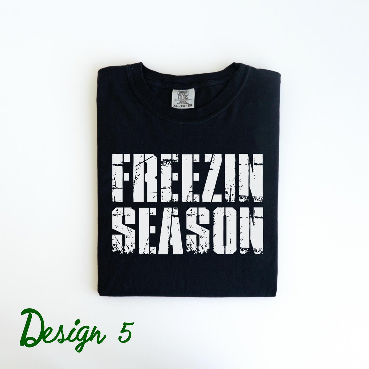 May include: Black t-shirt with the words "FREEZIN SEASON" in distressed white lettering. The shirt is folded neatly on a white surface. The words "Design 5" are written in green script in the lower left corner.