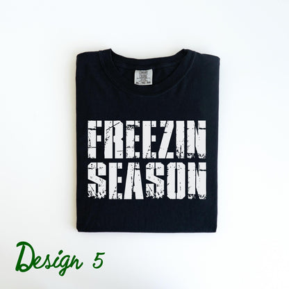 May include: Black t-shirt with the words "FREEZIN SEASON" in distressed white lettering. The shirt is folded neatly on a white surface. The words "Design 5" are written in green script in the lower left corner.