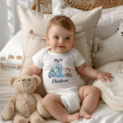 May include: A baby wearing a white onesie with the text "My 1st Christmas" and a graphic of a Christmas tree, penguins, and Santa Claus. The baby is sitting on a bed with pillows and a teddy bear.