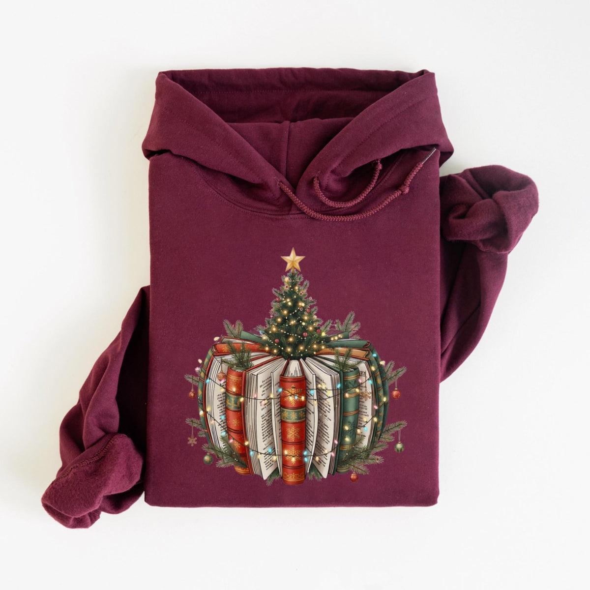May include: Burgundy hoodie featuring a Christmas tree design atop a stack of books. The tree is adorned with lights and a star, while the books are wrapped in festive lights and greenery. A cozy and festive apparel item.