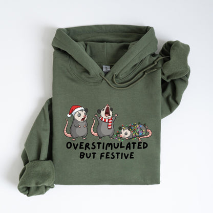 May include: Olive green hoodie featuring a graphic of three opossums. One wears a Santa hat, another a scarf, and the third is wrapped in Christmas lights. The text "OVERSTIMULATED BUT FESTIVE" is below the opossums.