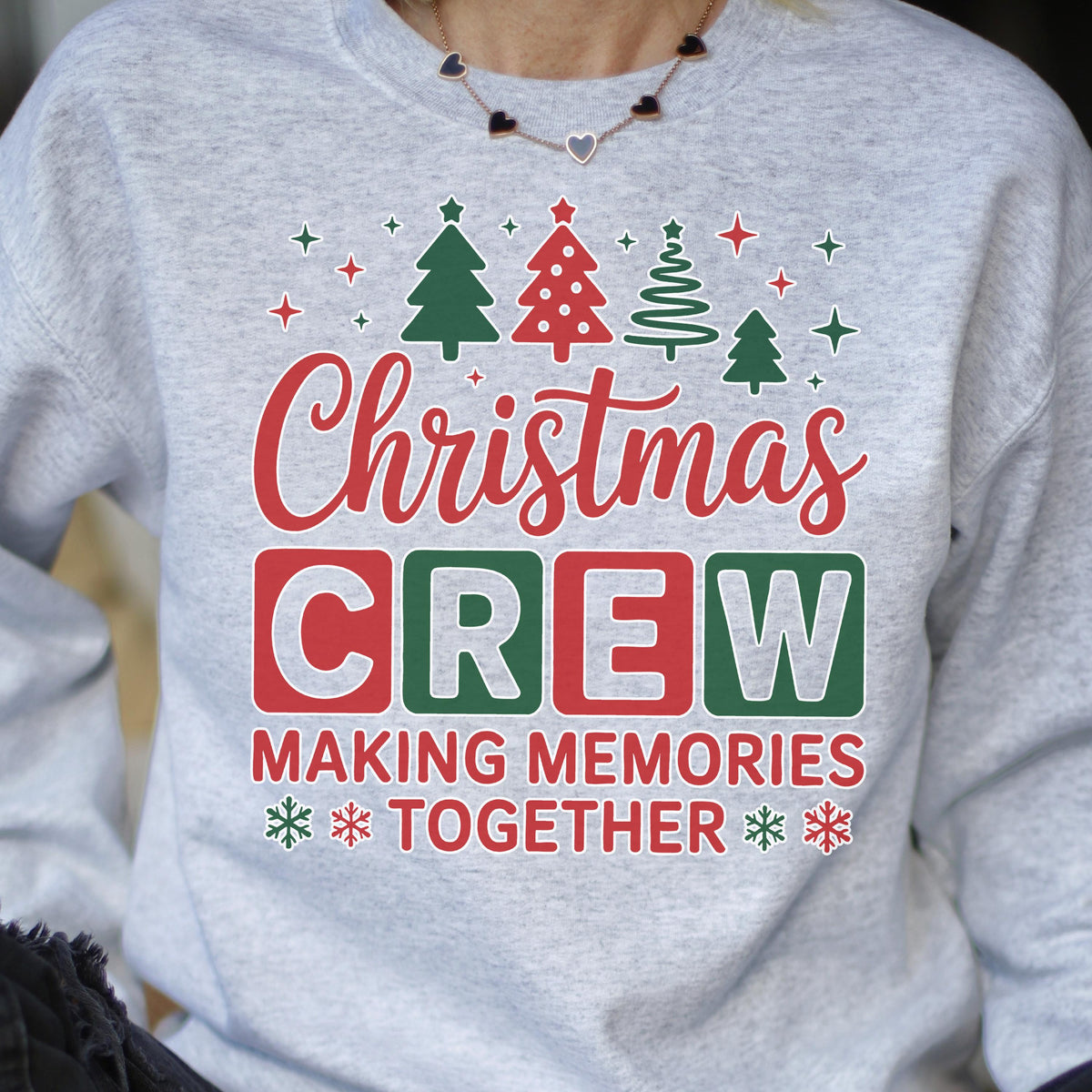 May include: A light gray crewneck sweatshirt featuring a festive graphic print. The design includes green and red Christmas trees, snowflakes, and the text "Christmas CREW MAKING MEMORIES TOGETHER" in a mix of red, green, and gray block lettering.