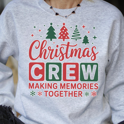 May include: A light gray crewneck sweatshirt featuring a festive graphic print. The design includes green and red Christmas trees, snowflakes, and the text "Christmas CREW MAKING MEMORIES TOGETHER" in a mix of red, green, and gray block lettering.