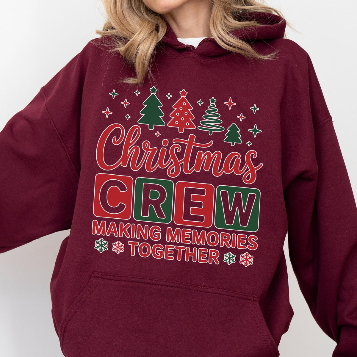 May include: A maroon hooded sweatshirt featuring a graphic print with the text Christmas CREW MAKING MEMORIES TOGETHER. The design includes red and green block letters, stylized Christmas trees, and small white snowflake icons.