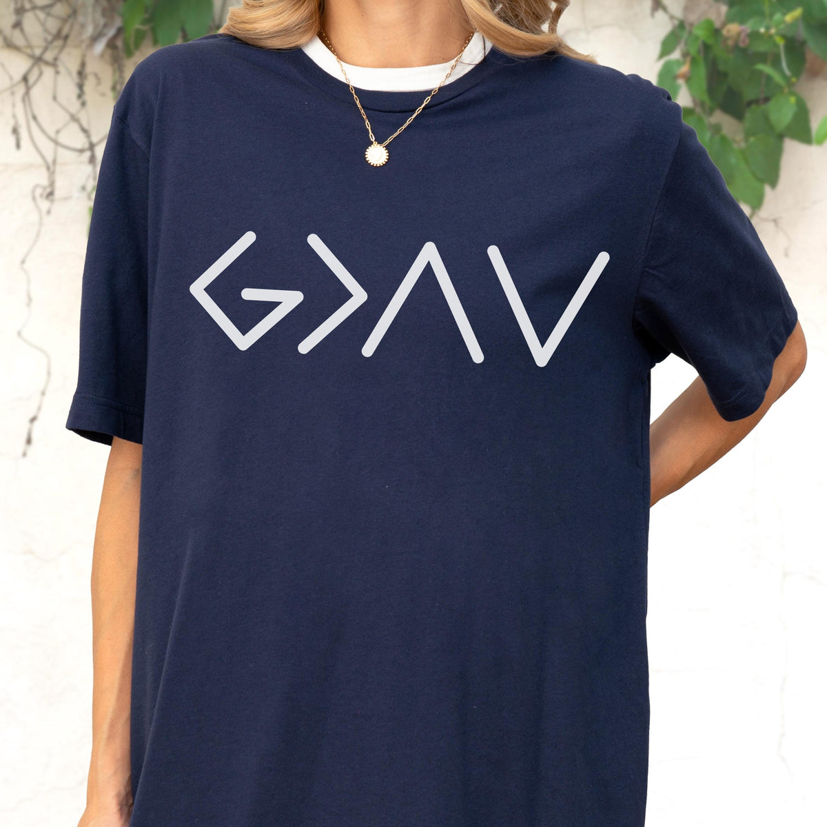 May include: A navy blue short-sleeved t-shirt featuring a white graphic print on the chest. The design consists of the mathematical symbols G > I V, which represent the phrase God is greater than the highs and lows. The shirt has a crew neckline.