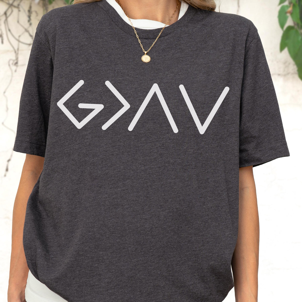 May include: A dark charcoal gray short-sleeve t-shirt featuring the white graphic text G > I V. The shirt has a crew neckline and a heathered fabric texture. A gold-toned chain necklace with a small circular pendant is worn around the neck.
