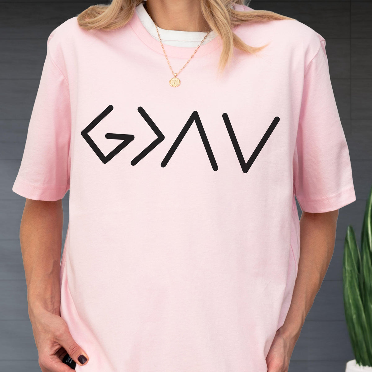 May include: A light pink short-sleeved t-shirt featuring the black graphic text G > I V centered on the chest. The shirt has a crew neckline and is worn over a white undershirt, paired with a thin gold chain necklace with a small circular pendant.