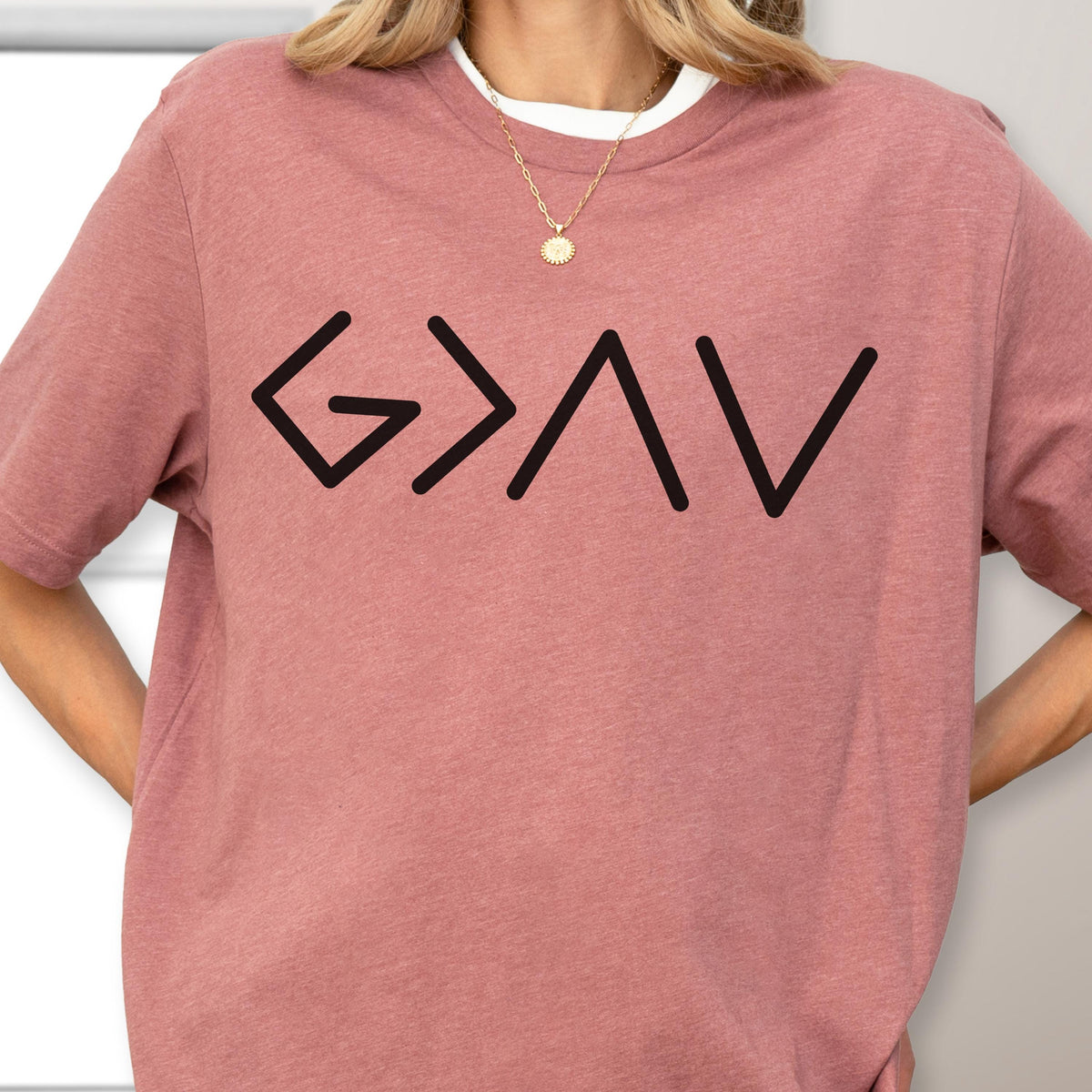 May include: A dusty rose short-sleeved t-shirt featuring a black graphic print of the symbols G > ^ v. The shirt has a crew neckline and is worn with a gold chain necklace featuring a small circular pendant resting against the collarbone area.
