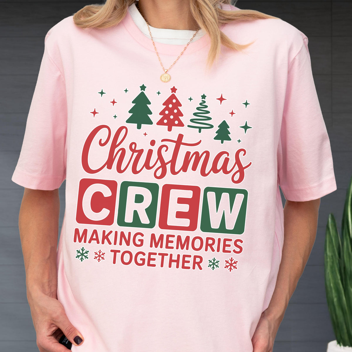 May include: A light pink short-sleeved t-shirt featuring a graphic print with the text Christmas CREW MAKING MEMORIES TOGETHER in red and green. The design includes illustrations of four stylized Christmas trees and small snowflakes.