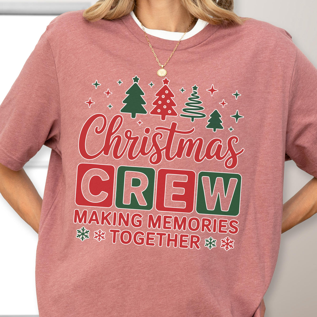 May include: A dusty rose short-sleeved t-shirt featuring a graphic print with the text Christmas CREW MAKING MEMORIES TOGETHER. The design includes red and green Christmas trees, stars, and snowflakes, with the word CREW in colored blocks.