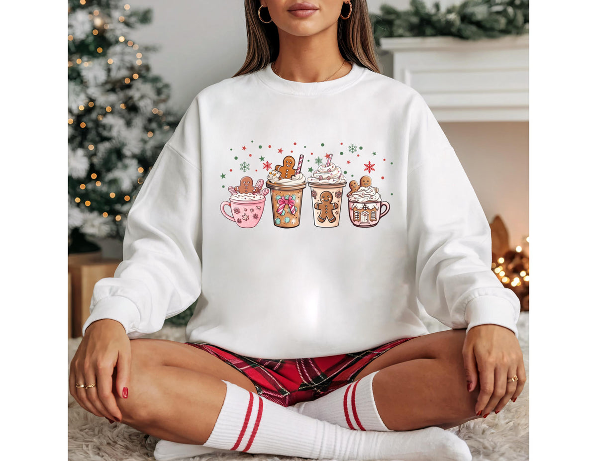 May include: White sweatshirt featuring a colorful graphic of four festive holiday drinks. Each drink is decorated with whipped cream, sprinkles, and gingerbread men. The design includes pink, brown, and green elements, perfect for the holiday season.