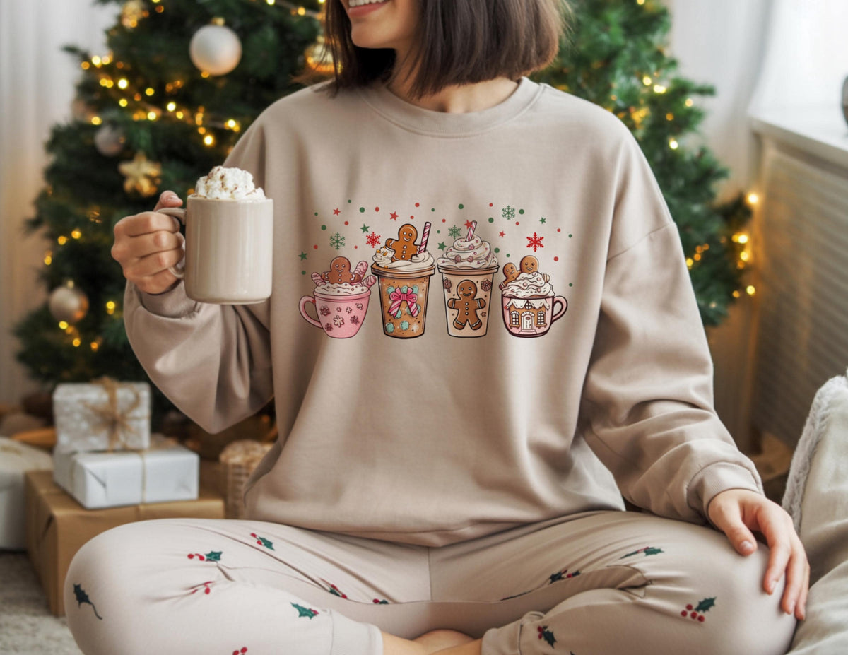 May include: A beige sweatshirt featuring a festive graphic of four gingerbread-themed drinks with whipped cream and gingerbread men. The sweatshirt is paired with matching pajama pants. A mug of whipped cream is held in the hand.