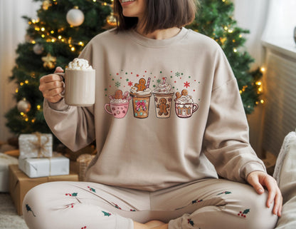 May include: A beige sweatshirt featuring a festive graphic of four gingerbread-themed drinks with whipped cream and gingerbread men. The sweatshirt is paired with matching pajama pants. A mug of whipped cream is held in the hand.