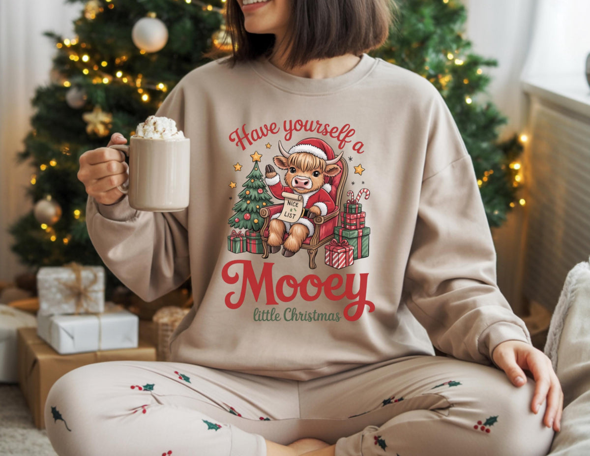 May include: Beige sweatshirt with a Christmas graphic featuring a cartoon cow in a Santa hat, a Christmas tree, and the text "Have yourself a Mooey little Christmas." The person is holding a mug.