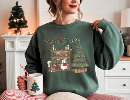 May include: A dark green sweatshirt featuring a Christmas-themed graphic. The graphic includes a Christmas tree, a bookshelf with festive decorations, and a ghost wearing a Santa hat. A white mug with a Christmas tree design is held in the hand.