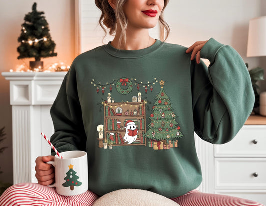 May include: A dark green sweatshirt featuring a Christmas-themed graphic. The graphic includes a Christmas tree, a bookshelf with festive decorations, and a ghost wearing a Santa hat. A white mug with a Christmas tree design is held in the hand.