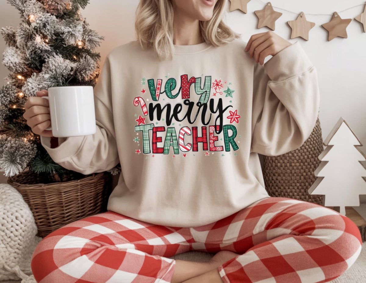 May include: A beige sweatshirt with the words "Very Merry Teacher" in a colorful, festive design. The person is wearing red and white plaid pants and holding a white mug. A Christmas tree and other holiday decorations are in the background.