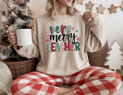 May include: A beige sweatshirt with the words "Very Merry Teacher" in a colorful, festive design. The person is wearing red and white plaid pants and holding a white mug. A Christmas tree and other holiday decorations are in the background.
