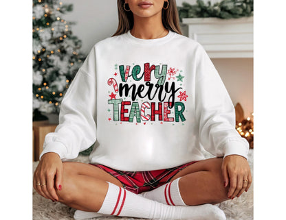 May include: White sweatshirt with the text "Very Merry Teacher" in a festive design. The text features red, green, and white colors with candy cane and snowflake accents. The sweatshirt is paired with red plaid shorts and white socks.