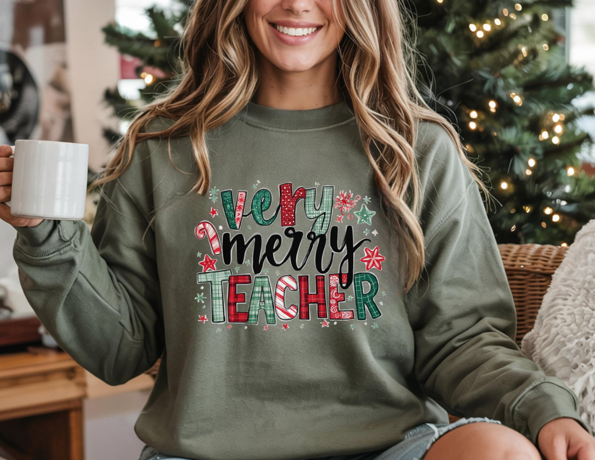 May include: An olive green sweatshirt with the text "Very Merry Teacher" in a festive design. The text incorporates red, green, and white colors with candy cane and star accents. The sweatshirt is long-sleeved.
