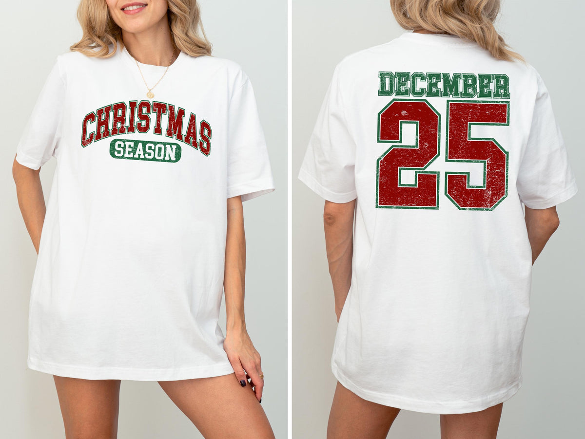May include: White t-shirt with "CHRISTMAS SEASON" in red and green arched text on the front. The back of the shirt displays "DECEMBER 25" in red and green block letters. The shirt has a vintage, distressed look.
