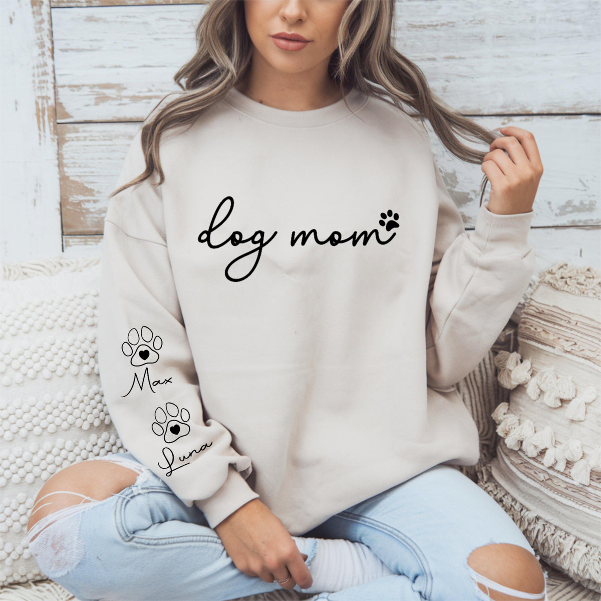May include: A beige sweatshirt with the words "dog mom" in black script and a paw print. The sleeve features two paw prints with the names "Max" and "Luna" below them. The sweatshirt is a casual style.