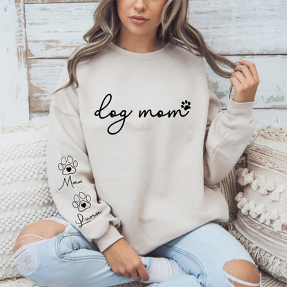 May include: A beige sweatshirt with the words "dog mom" in black script and a paw print. The sleeve features two paw prints with the names "Max" and "Luna" below them. The sweatshirt is a casual style.