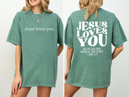May include: A sage green t-shirt with the text "Jesus loves you" on the front. The back of the shirt features the words "JESUS LOVES YOU" in a wavy font, along with the Bible verse 1 John 4:19.