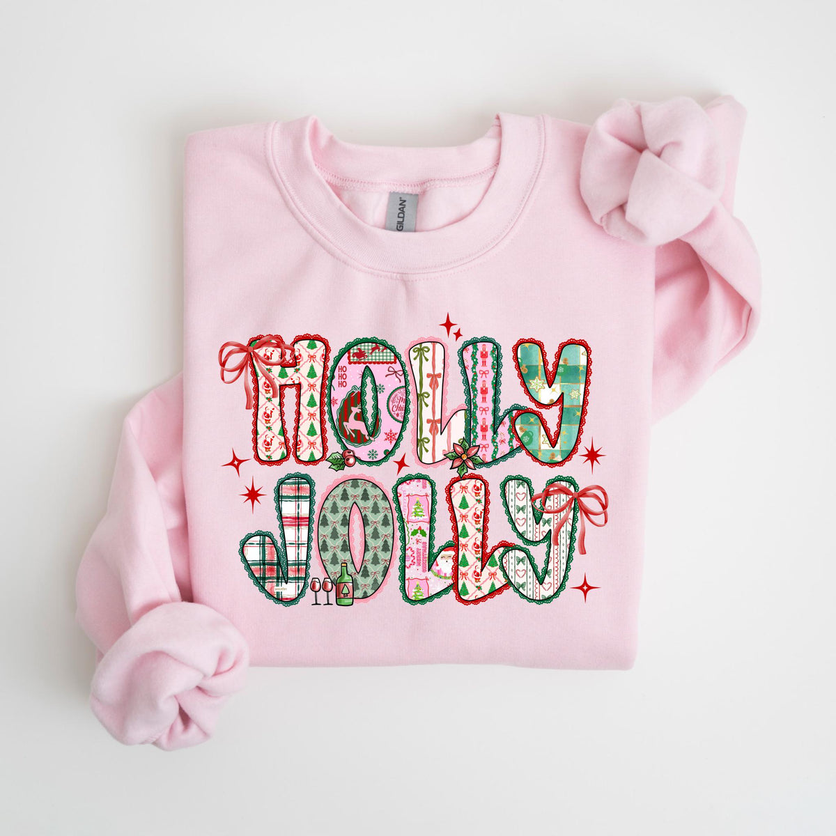 May include: A light pink sweatshirt with the words "HOLLY JOLLY" in a patchwork design. The letters are filled with Christmas-themed patterns, including plaid, trees, and ornaments. Red bows and stars accent the design.
