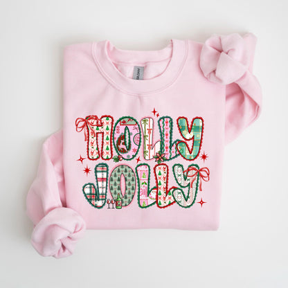 May include: A light pink sweatshirt with the words "HOLLY JOLLY" in a patchwork design. The letters are filled with Christmas-themed patterns, including plaid, trees, and ornaments. Red bows and stars accent the design.
