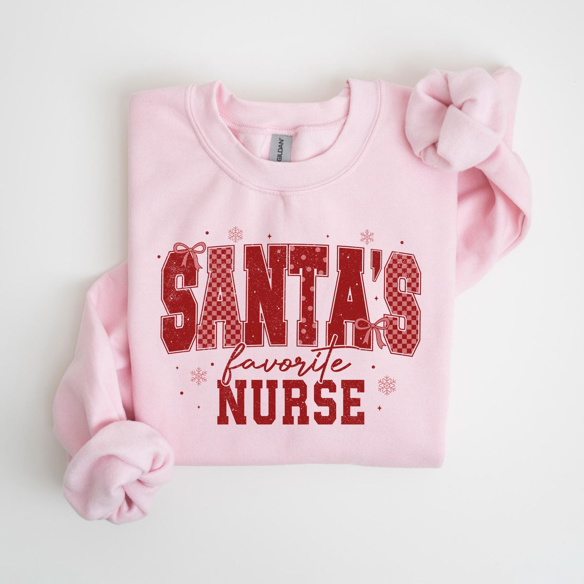May include: A light pink sweatshirt with the text "SANTA'S favorite NURSE" in red. The text has a checkered pattern and is decorated with bows and snowflakes. The sweatshirt is a crew neck style.