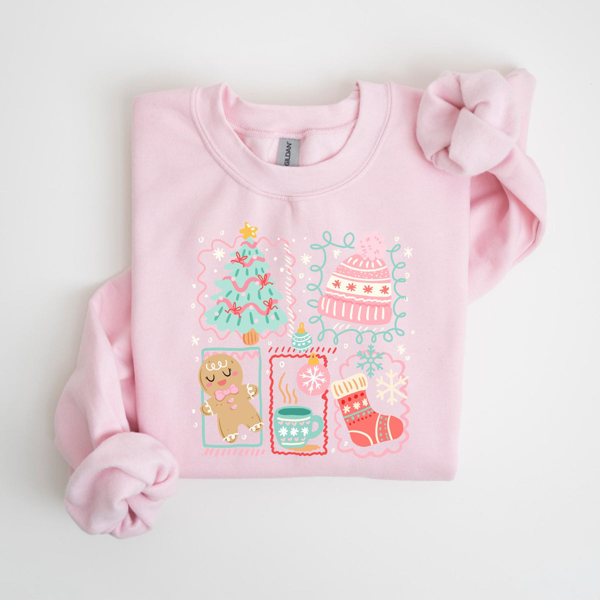 May include: A light pink sweatshirt with a festive graphic. The graphic includes a Christmas tree, a gingerbread man, a mug of hot cocoa, a stocking, and a winter hat, all in a whimsical style. The design is outlined with a light blue border.