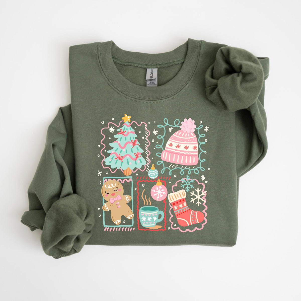 May include: Olive green sweatshirt with a festive Christmas design. The graphic includes a Christmas tree, a pink hat, a gingerbread man, a mug of hot cocoa, and a Christmas stocking. The design is outlined in a pink and white border.